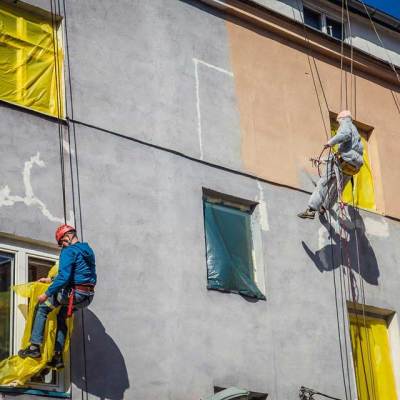 wysokościowe prace malarskie, pracownicy zabezpieczają folią okna i wykonują hydrodynamiczne malowanie elewacji